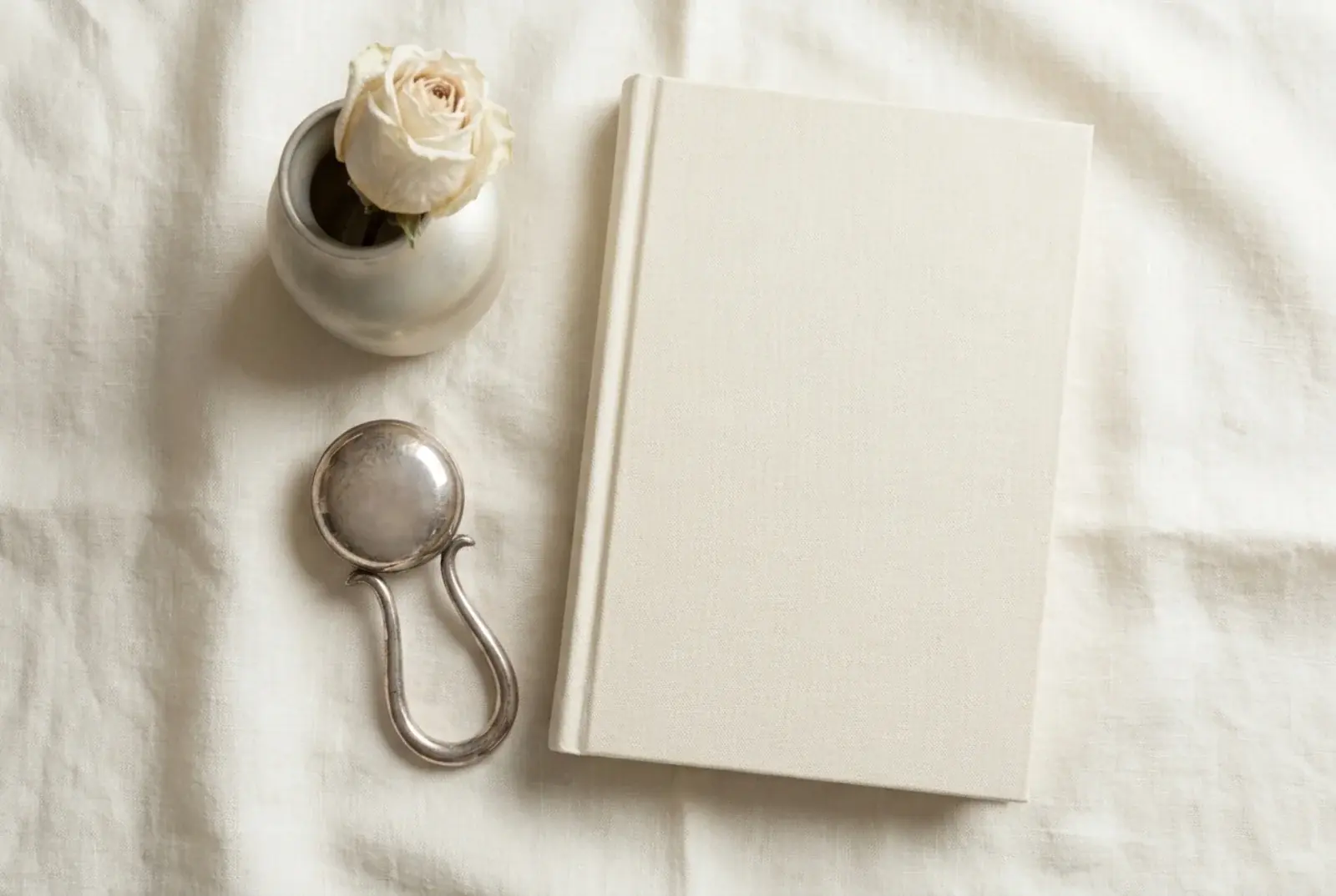 A small silver christening cup on a white linen runner beside a matte hardcover book and a folded white knitted blanket in soft window light against pale wood.