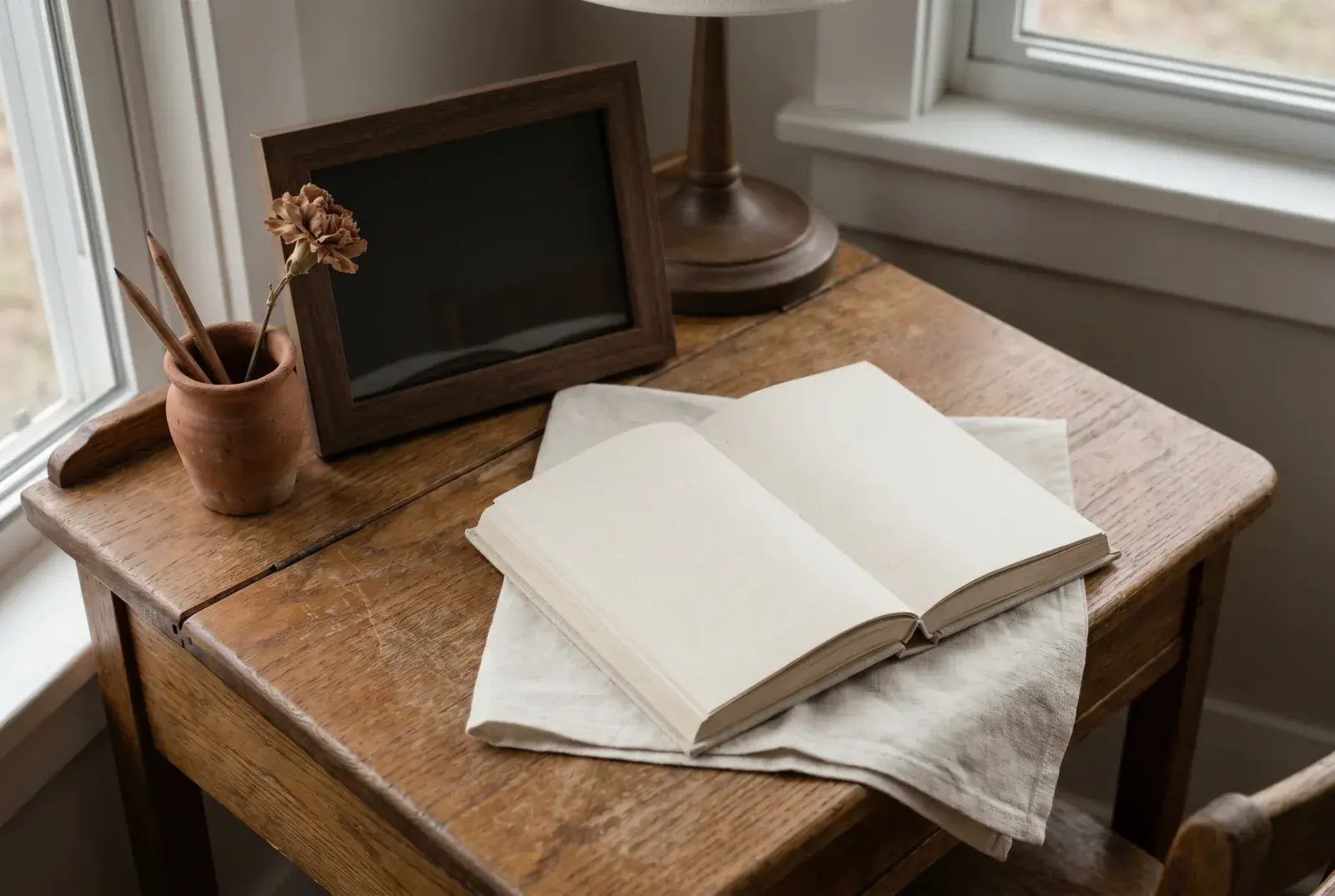 Oak desk still life: an open hardcover storybook on cream linen beside an empty wooden picture frame and a small clay vessel with two pencils — book and absence.