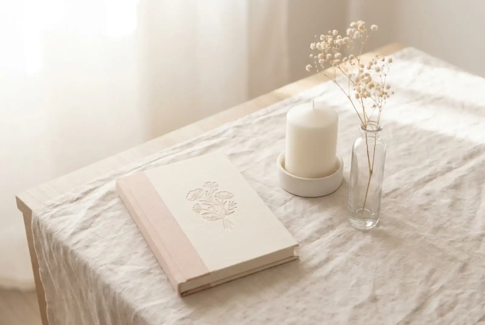 A matte hardcover book on a low oak table beside a single white candle in a ceramic holder, a folded muslin blanket, and a small wooden figure in afternoon light.