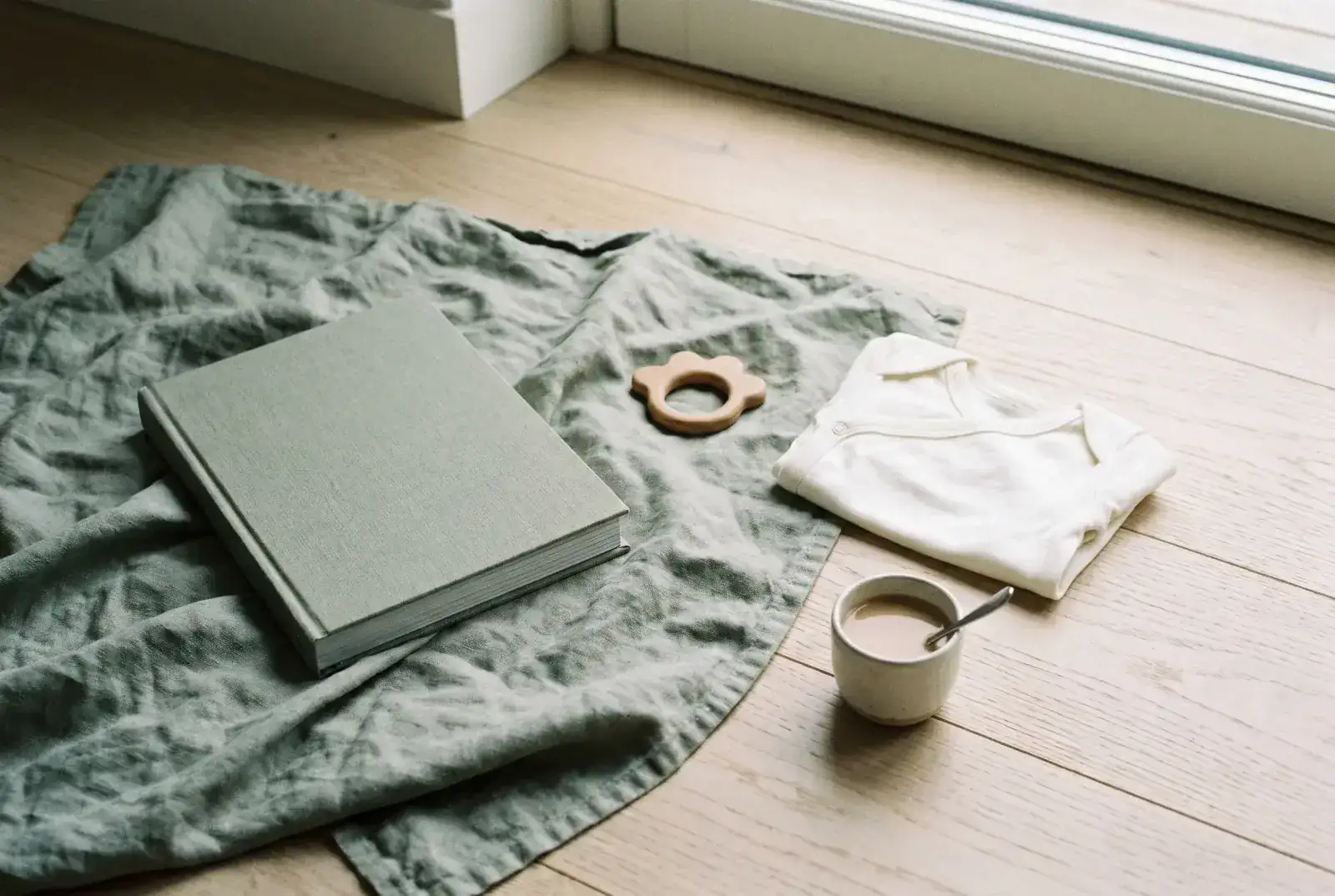 A hardcover book on a sage linen blanket beside a small wooden teether, a folded white onesie, and a ceramic cup of milky tea on a pale oak floor.
