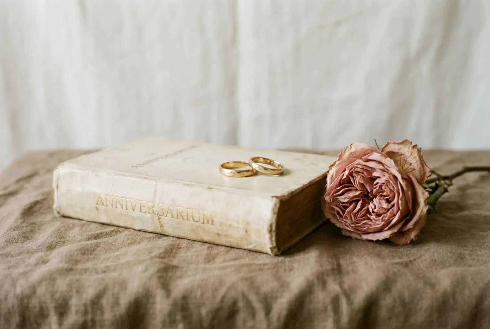 A matte-printed hardcover golden anniversary keepsake book and a small pair of gold rings resting on warm linen in soft window light.
