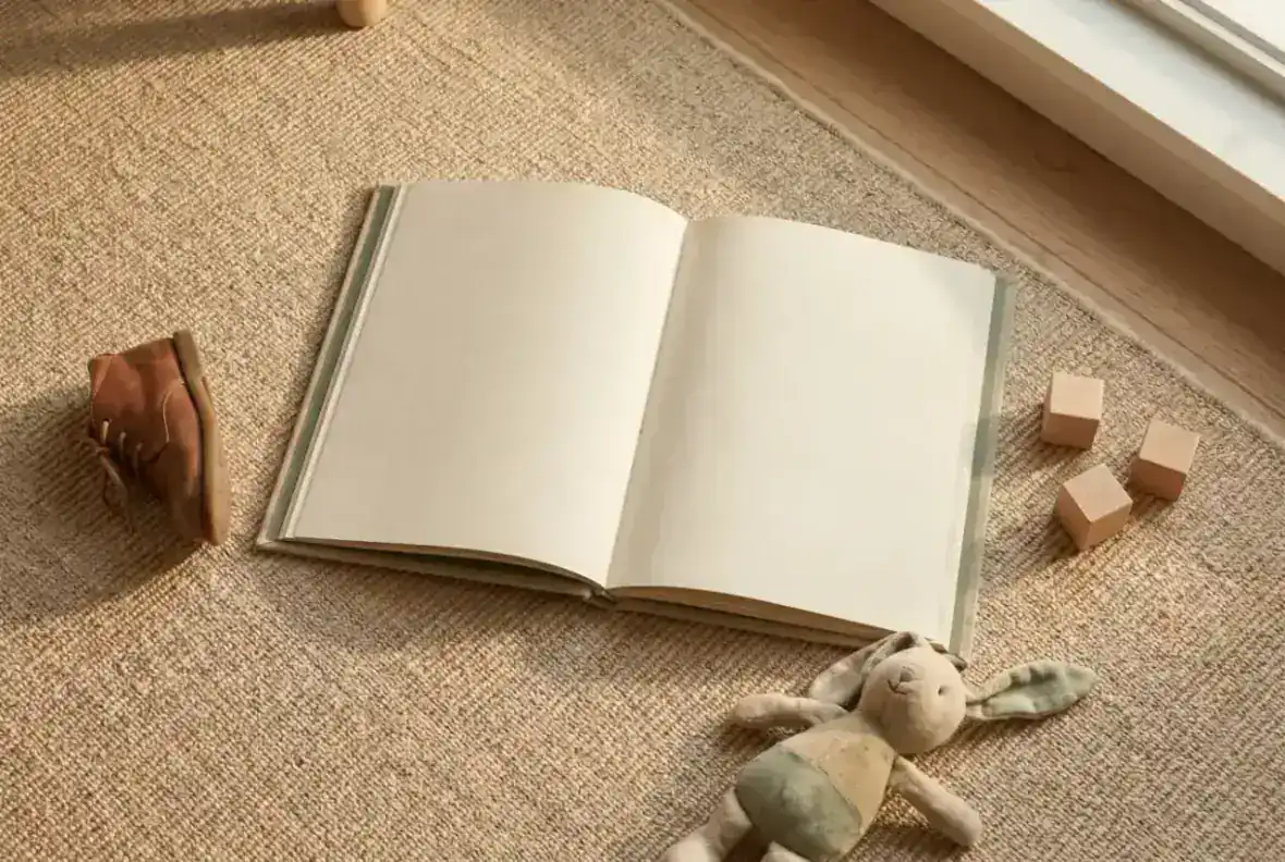 A hardcover storybook open at the center of a sisal rug beside scattered wooden blocks, a small leather shoe, and a soft fabric bunny — the book holds the room.
