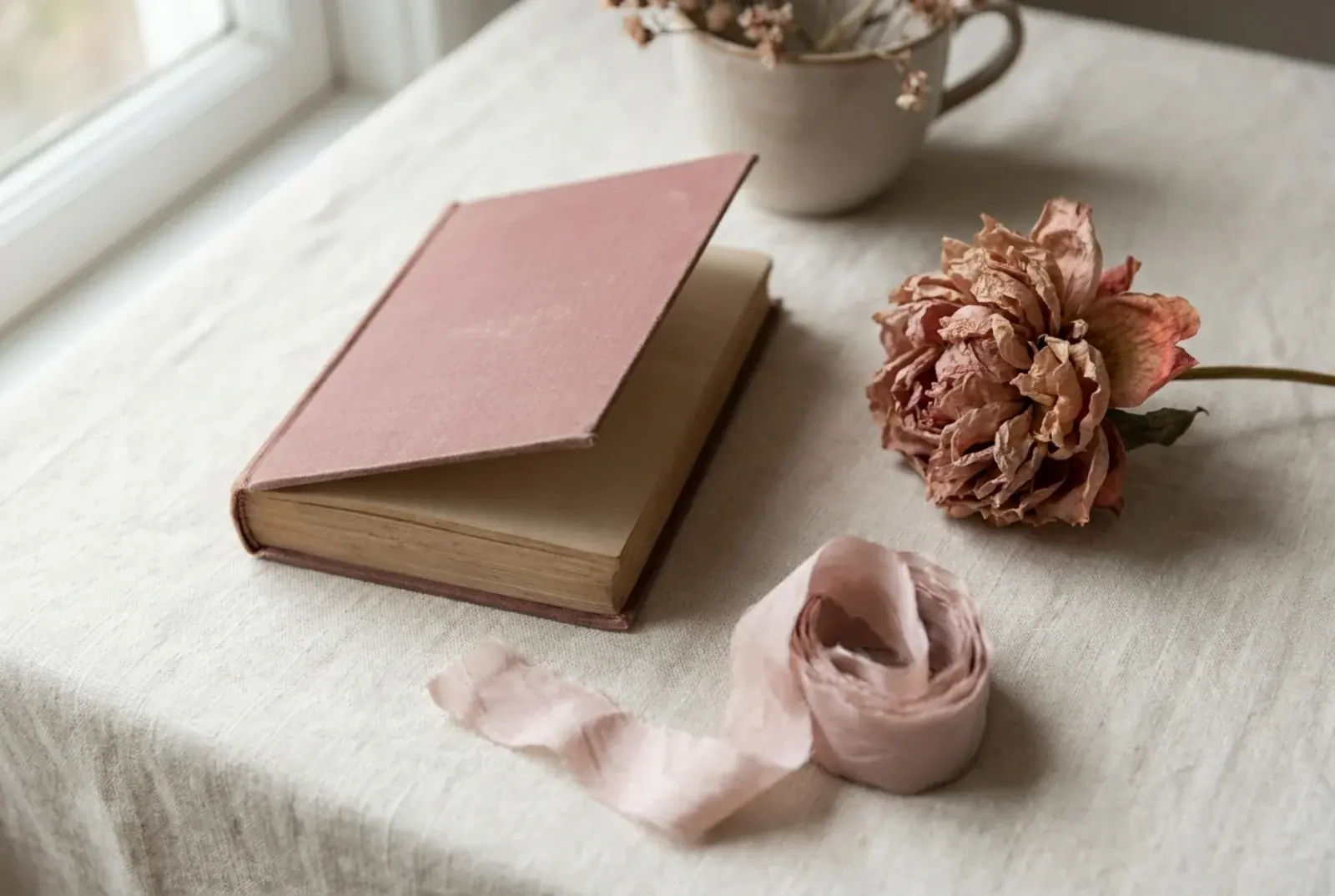 A matte hardcover book on a pale wooden child's chair beside a folded pink linen pinafore and a small wooden Maileg mouse in warm afternoon window light.