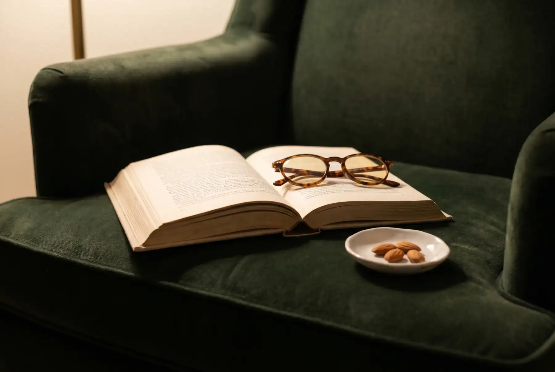 A cream hardcover book lying open on a forest-green velvet armchair, tortoiseshell reading glasses on the right page, a porcelain dish of almonds alongside, warm lamp light.