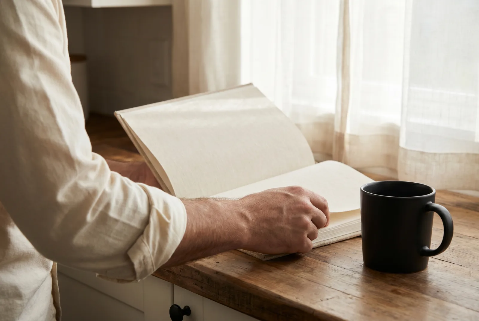 A hardcover book open to a blank page held against a man's forearm in a rolled cream shirt cuff on a kitchen counter beside a matte black coffee mug.