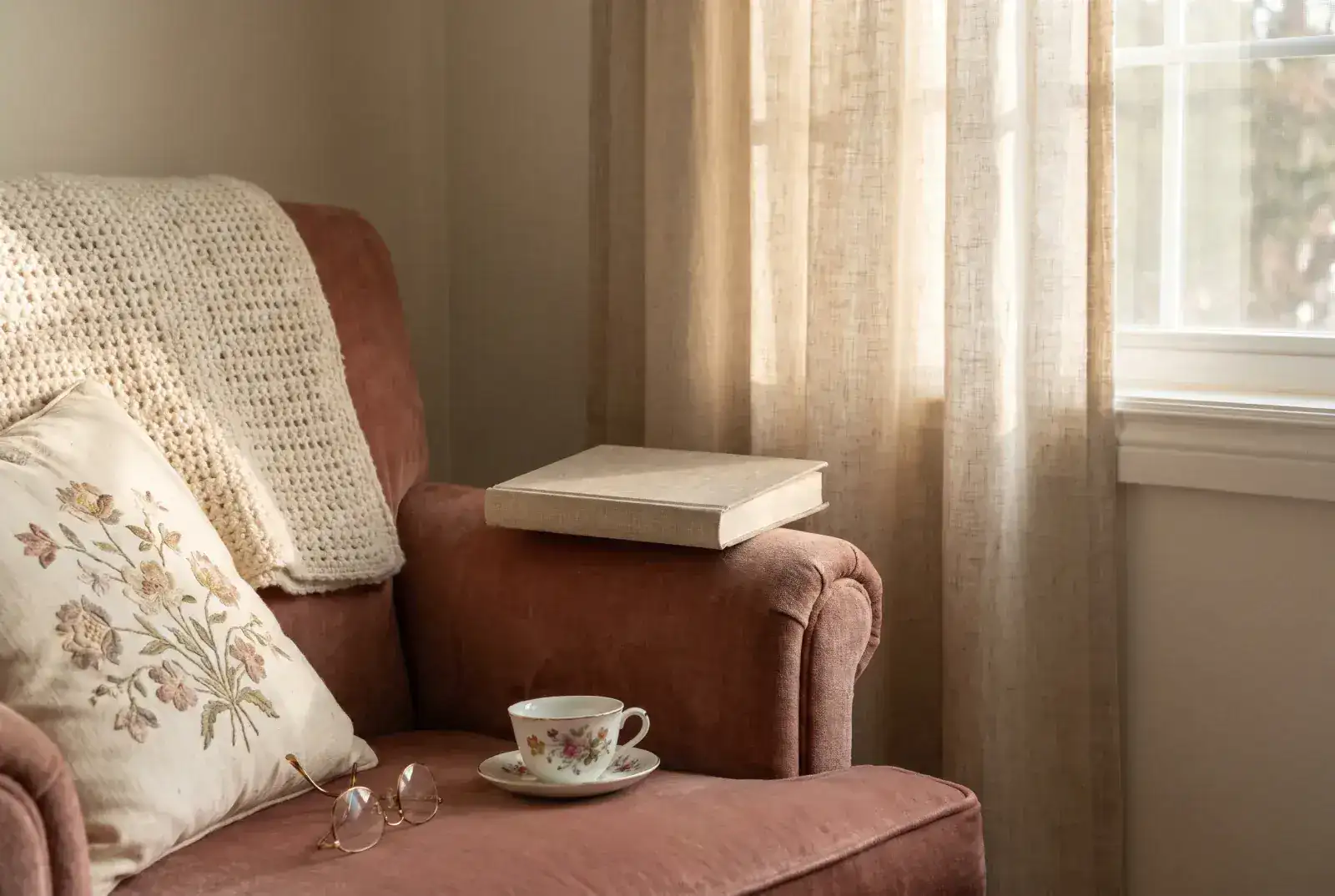 A hardcover book on a velvet chair arm beside a porcelain teacup, reading glasses folded on an embroidered cushion, and a crocheted cream blanket on the chair back.