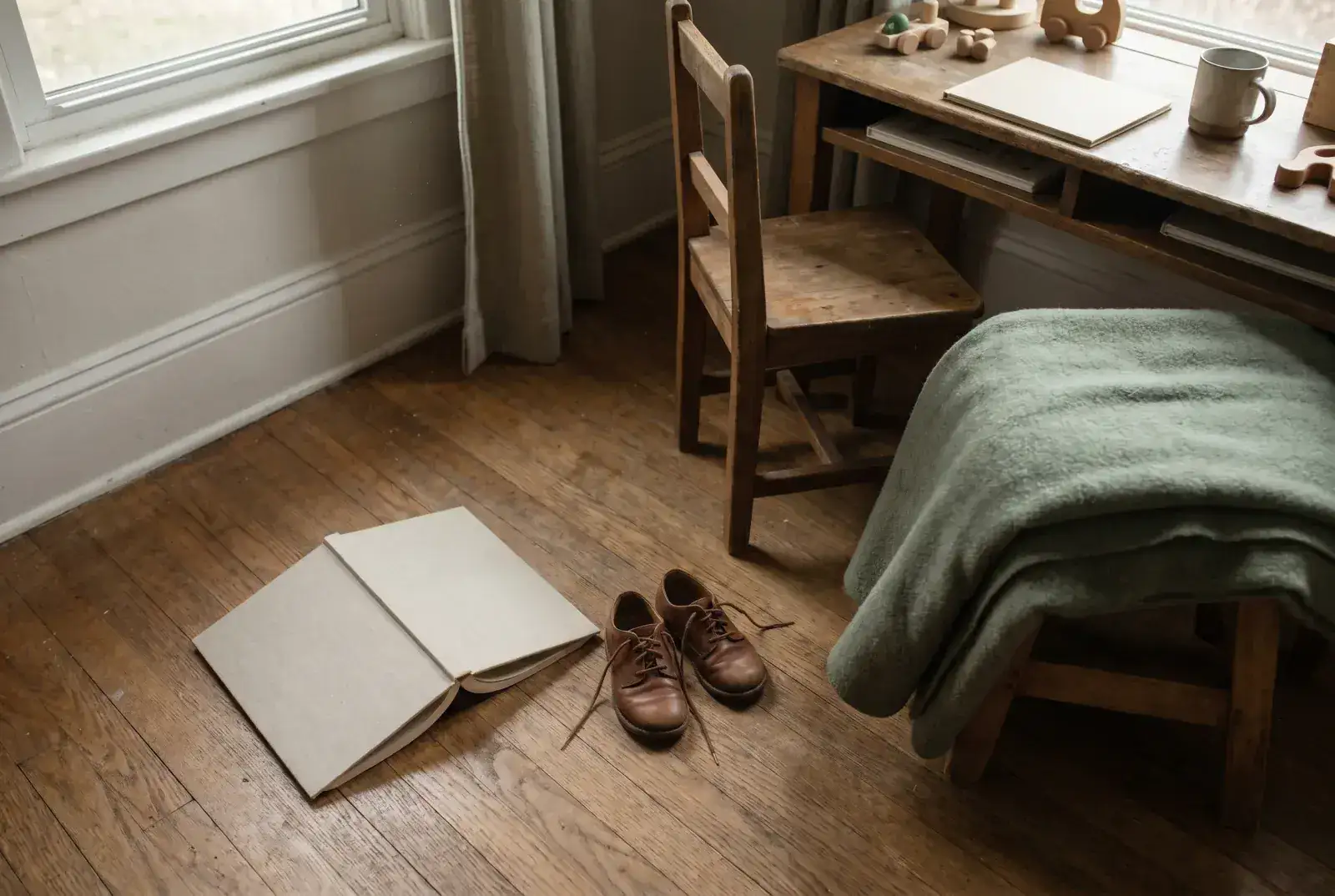A single hardcover storybook resting on an oak floor in soft sunlight beside a small pair of leather shoes and a folded sage wool blanket — quiet pause, no devices.
