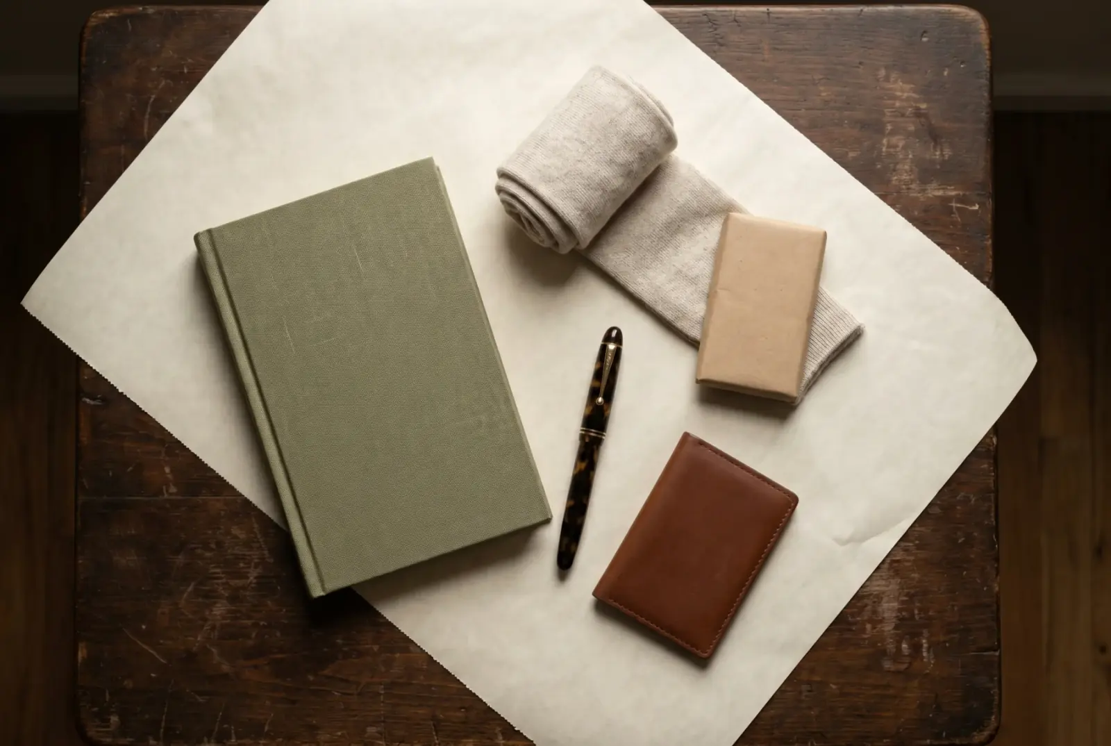 Curated small objects on cream paper: a clothbound notebook, rolled wool socks, an artisan chocolate bar, a fountain pen, and a leather card case in soft natural light.