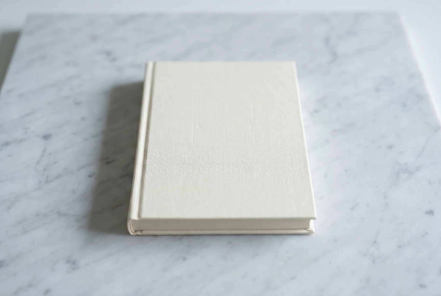 A single cream hardcover book resting on a pale Carrara marble surface with large negative space around it, faint shadow, cool diffuse light, museum-still composition.