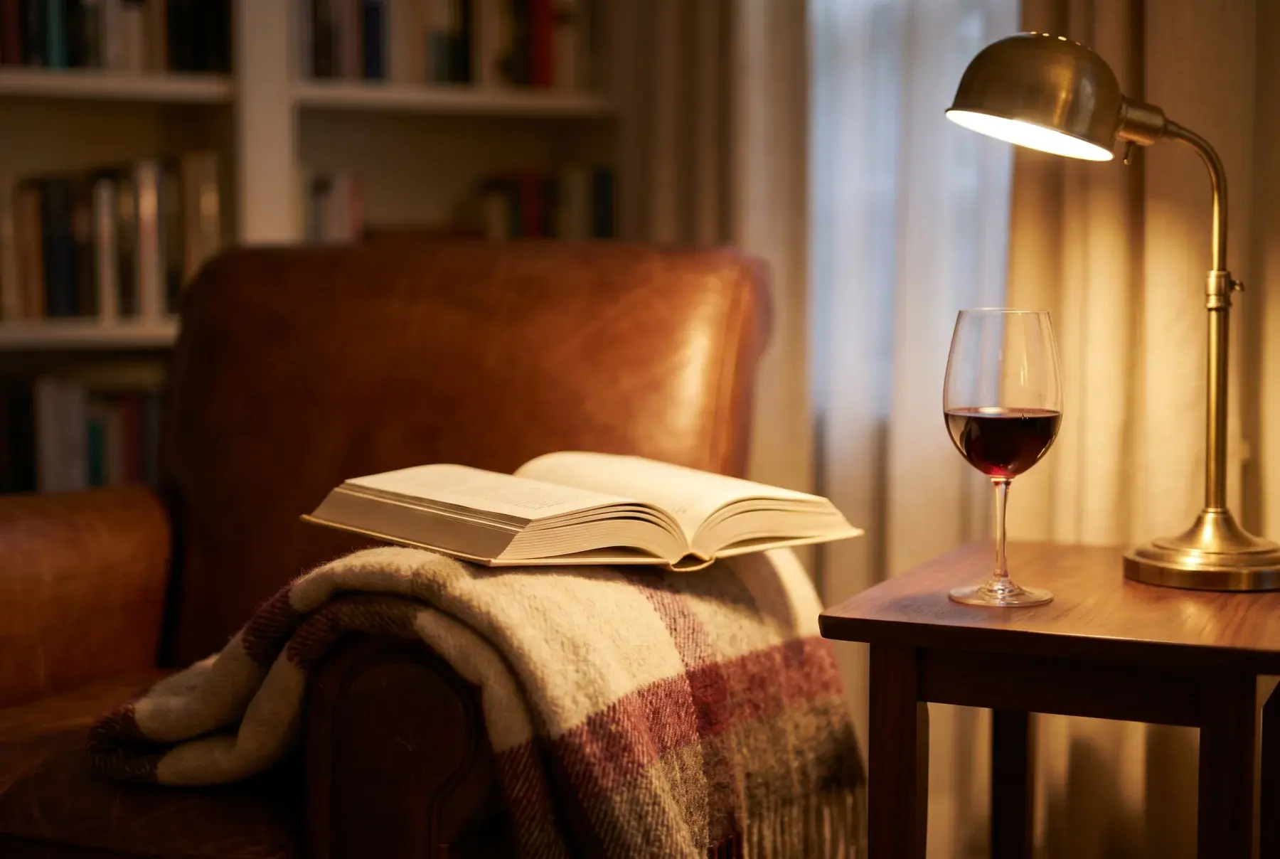 An open hardcover book on a wool blanket over a worn leather armchair, with a half-finished glass of red wine and a brass lamp casting warm light.