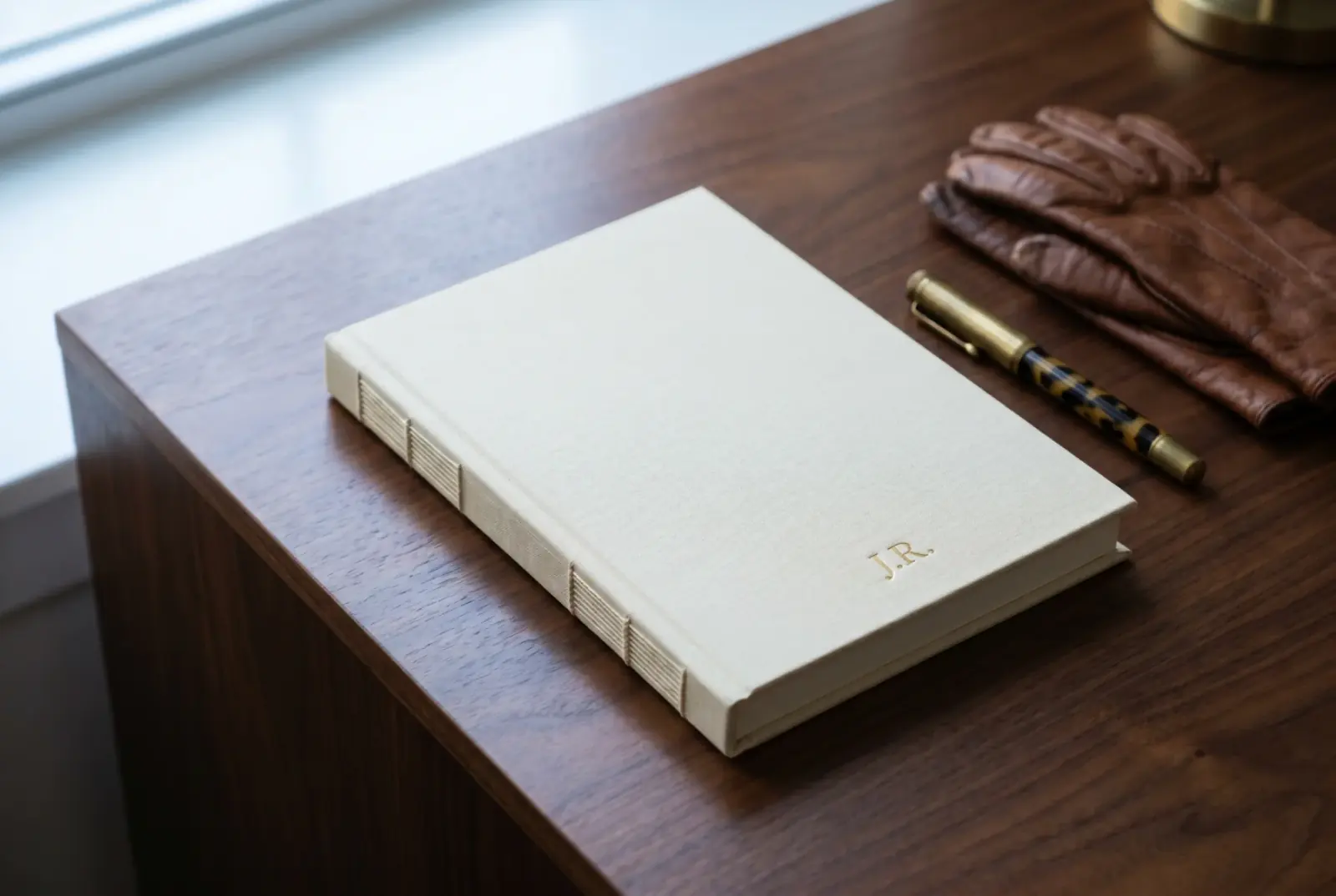 A cream hardcover book lying flat on a dark walnut writing desk, with leather gloves and an antique fountain pen alongside, north-window light falling across the cover.