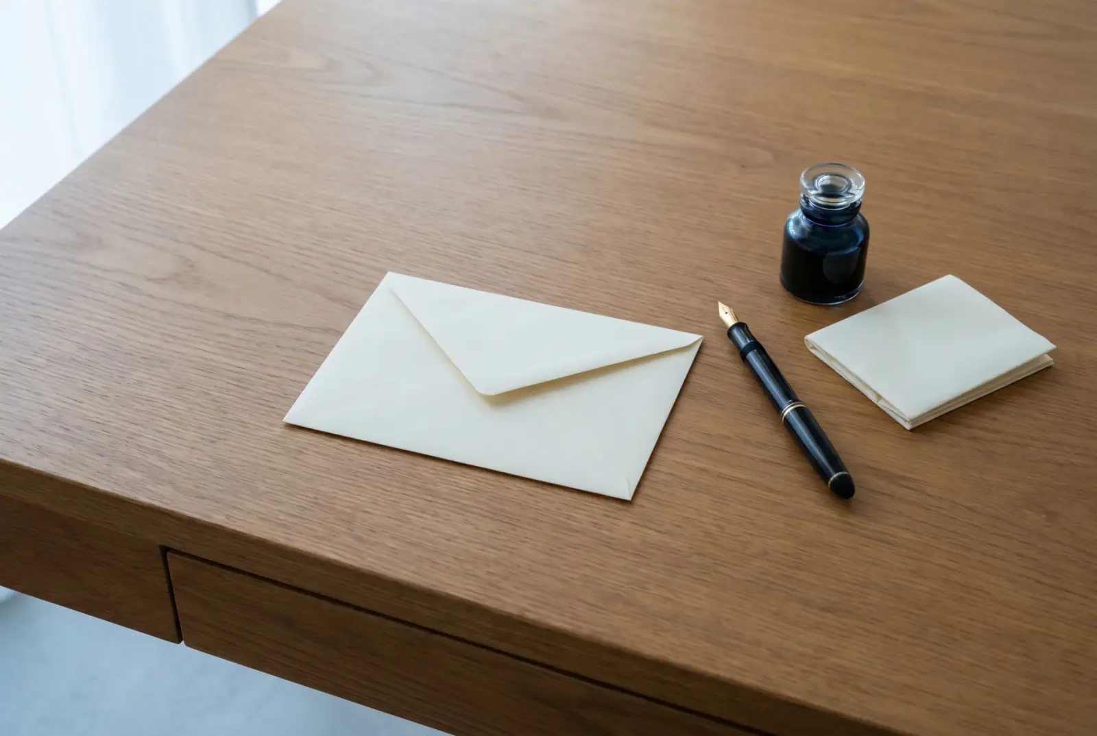 A sealed cream envelope resting on a polished oak desk beside a vintage fountain pen, a small glass inkwell, and a sheet of cream blotting paper, with soft north-window light.